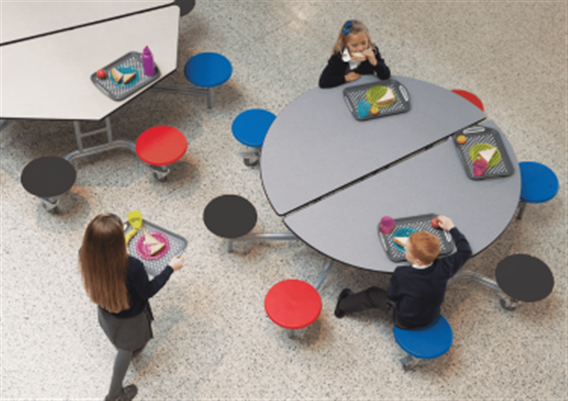 A child is eating their lunch on a circular table, with a grey blue top and 3 different coloured seats (red, black and blue). A Girl is walking over to the table.